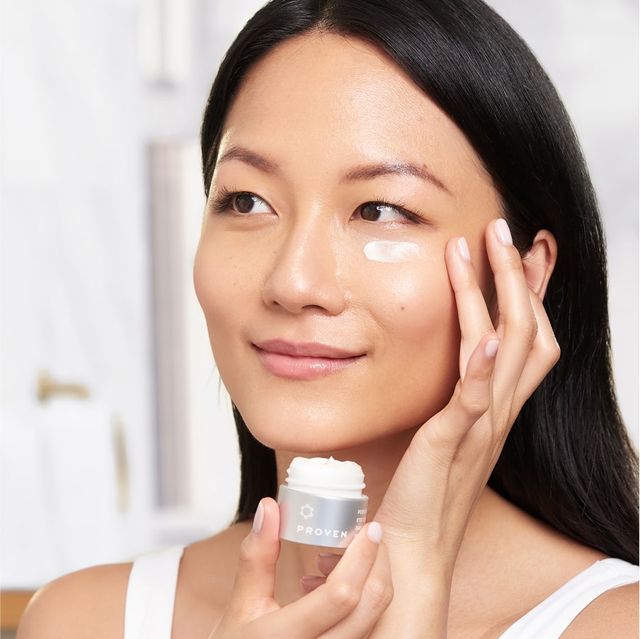 A woman patting the day eye cream under her eye, the open jar in her other hand.